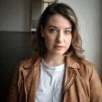Headshot of Jil Clesse, Physical Lab associate teaching artist, wearing a white t-shirt and brown leather jacket