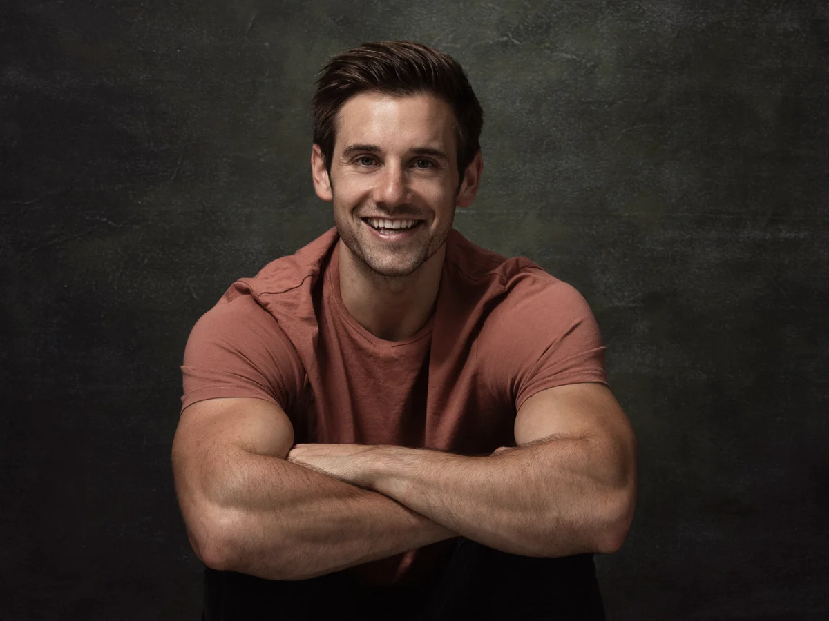 Alex Crockford sitting with arms crossed against dark background and smiling