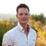 Ed Pike in a white button-down shirt standing with his arms behind his back against the wild hills of Crete.