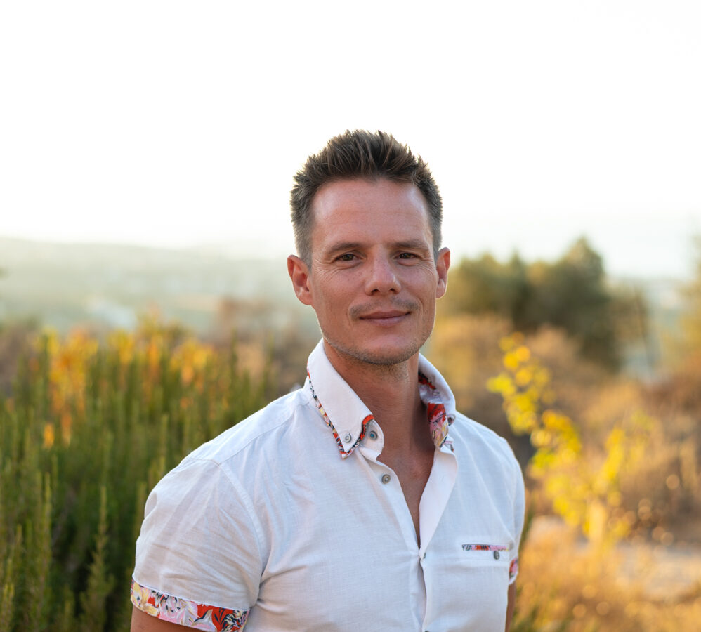 Ed Pike in a white button-down shirt standing with his arms behind his back against the wild hills of Crete.