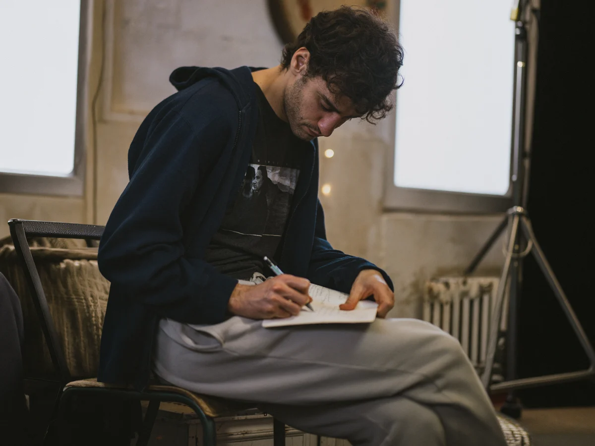 Young man with dark hair sitting on a chair and writing in a notebook.