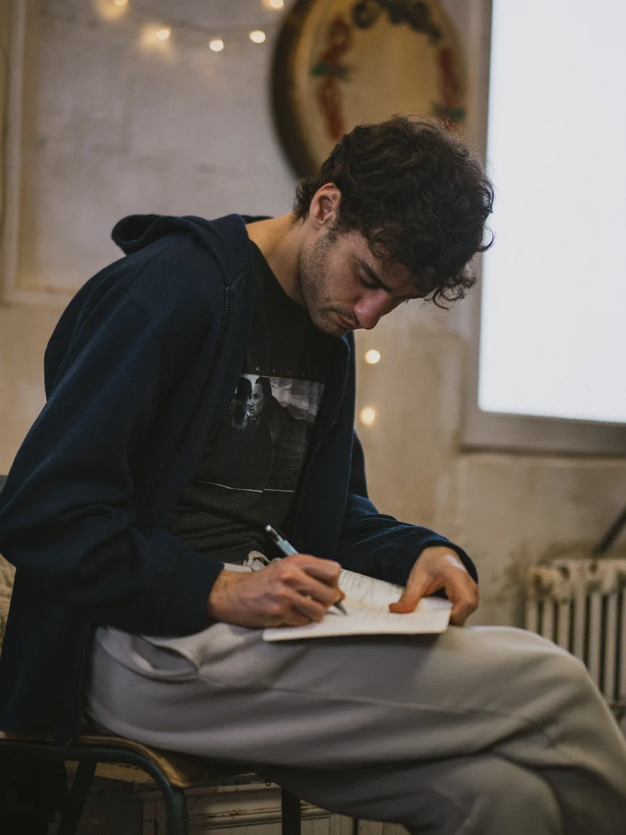Young man with dark hair sitting on a chair and writing in a notebook.